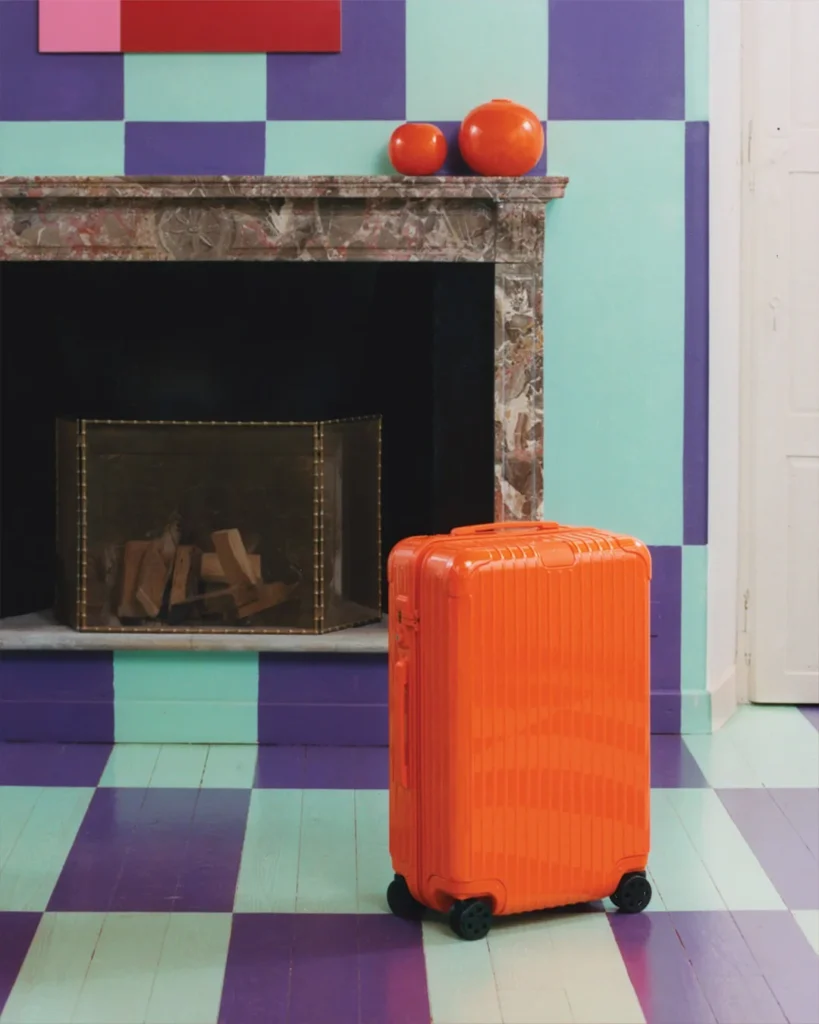 The iconic orange RIMOWA Essential suitcase in front of a stylish fireplace with a retro wallpaper.