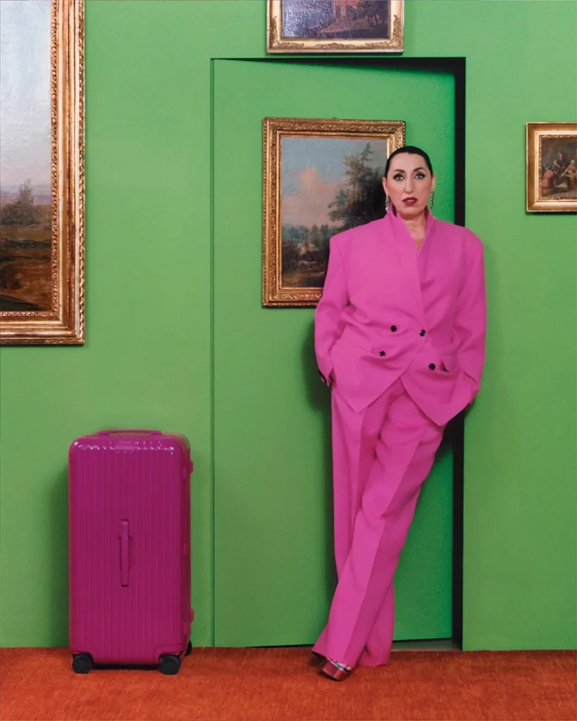 Rossy de Palma and a magenta RIMOWA suitcase in front of a green wall of paintings.