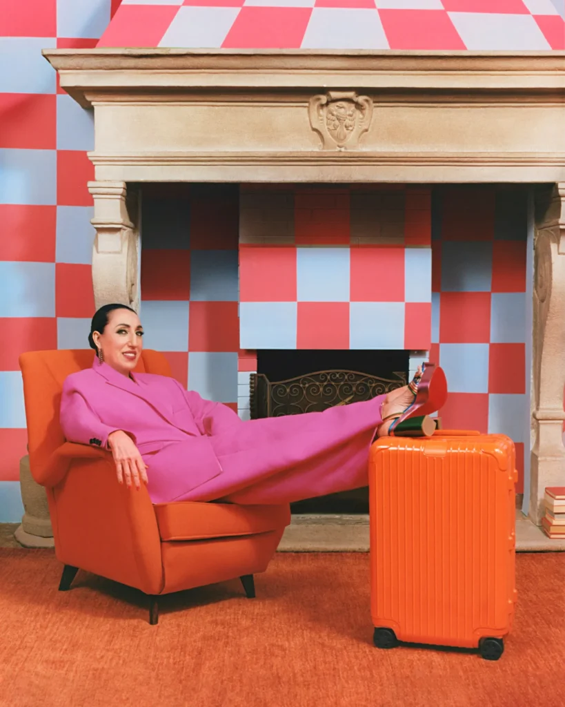 Actress Rossy de Palma in a pink costume with an orange RIMOWA suitcase.