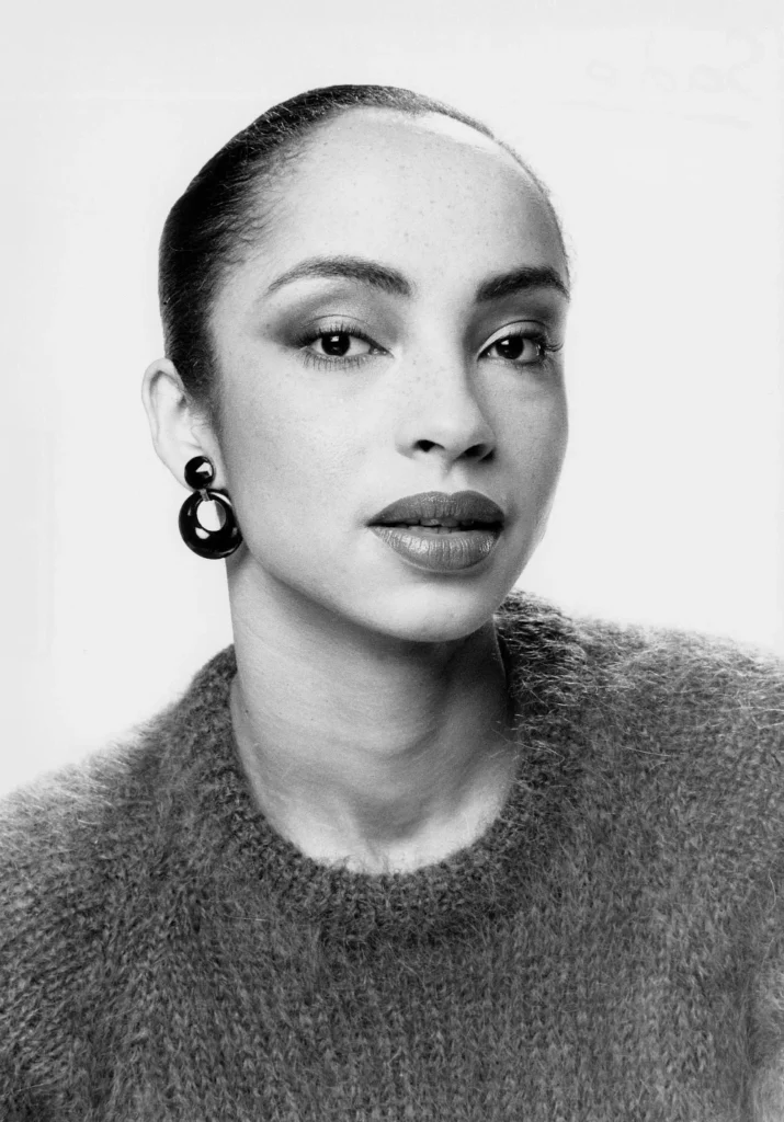 Black and white portrait photo of Sade Adu in a knitted sweater with massive earrings.