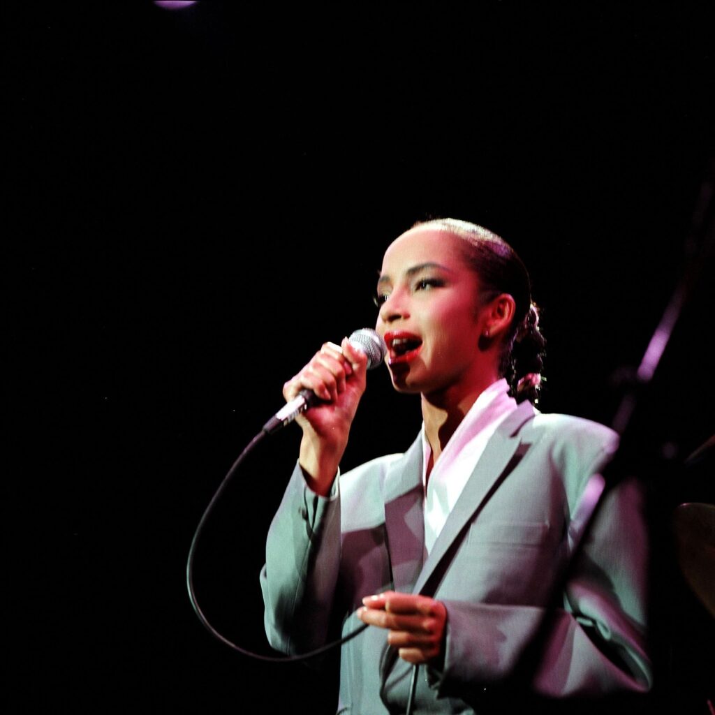 Sade Adu performing live on stage in a sharp grey suit.