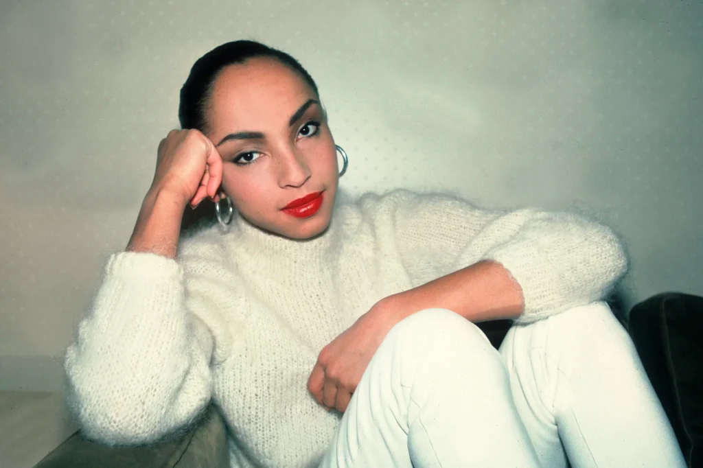 Minimalist portrait of Sade Adu in a white turtleneck with silver hoop earrings.