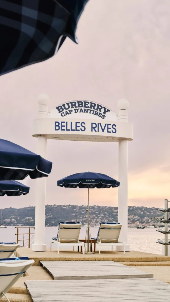 Beach pier with the iconic Burberry Cap d'Antibes Belles Rives sign, navy blue umbrellas, and sunbeds during a scenic sunset.