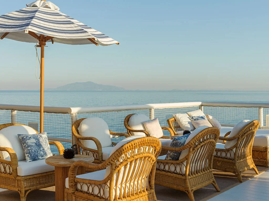 Terrace of the Dior beach club on Capri island with wicker armchairs, blue and white Toile de Jouy cushions, and an open sea view.