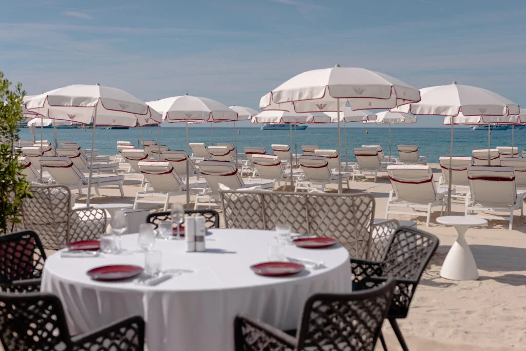 Luxury sandy beach in Cannes featuring white umbrellas and sunbeds with red trim by Emporio Armani, with a set dining table in the foreground.
