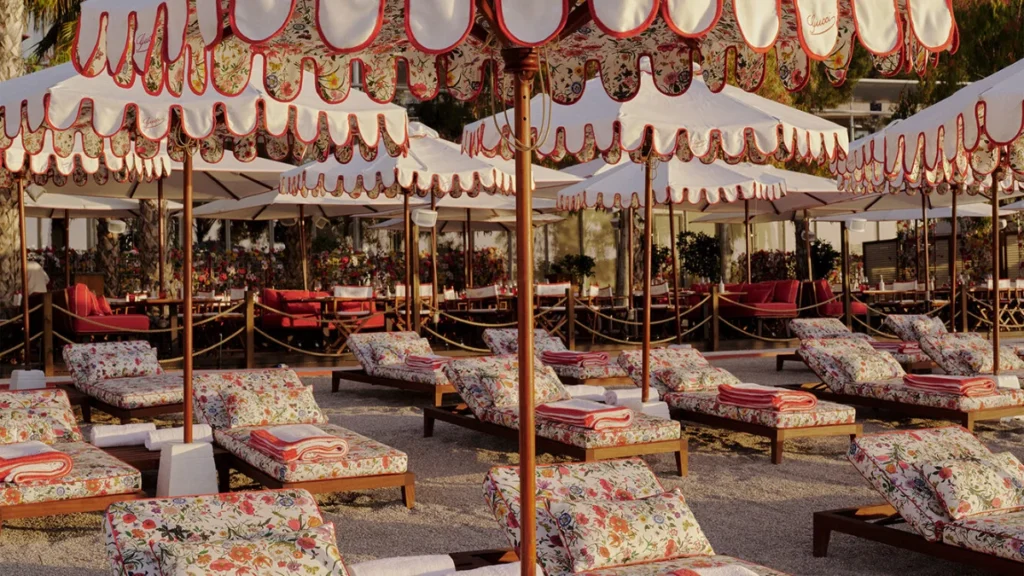 Beach sunbeds and ruffled umbrellas featuring the iconic Gucci Flora floral print with red trim at a luxury seaside resort.