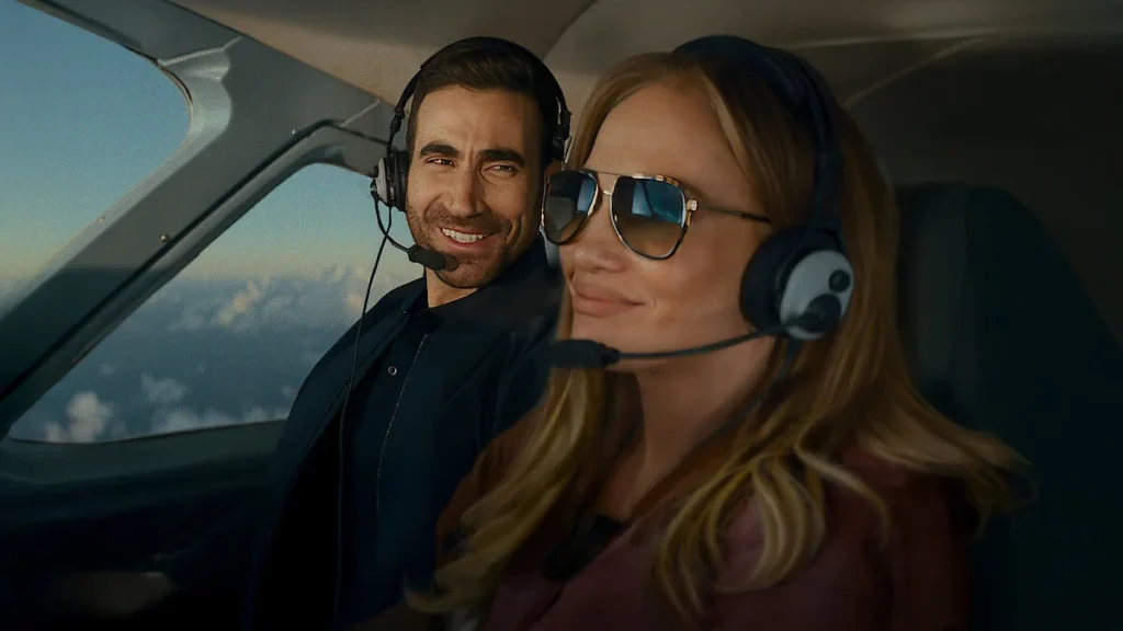 Jennifer Lopez wearing sunglasses and Brett Goldstein with aviation headsets in an airplane cockpit in the movie Office Romance.