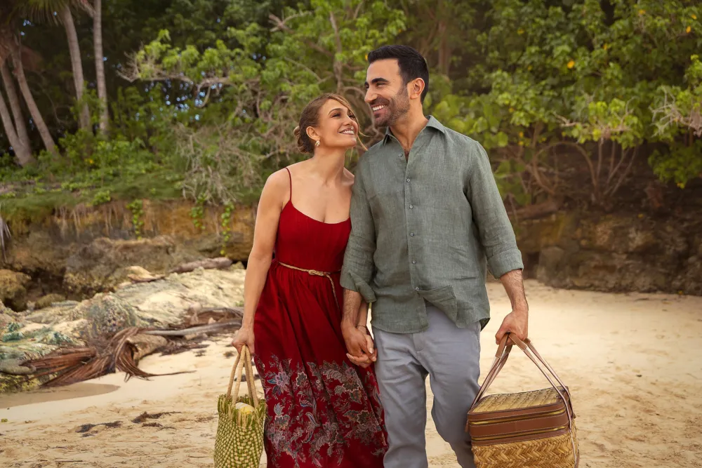 Jennifer Lopez in a red dress and Brett Goldstein holding hands while walking on the beach in the movie Office Romance.