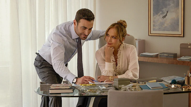 Brett Goldstein leaning over the desk where Jennifer Lopez is sitting in a scene from the movie Office Romance.