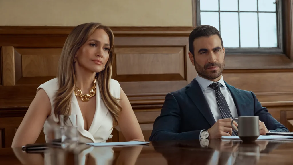 Jennifer Lopez and Brett Goldstein sitting next to each other at a large table in a meeting room in the movie Office Romance.