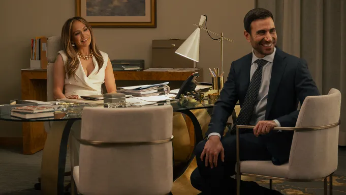 A smiling Jennifer Lopez sitting at her desk in her office across from Brett Goldstein in the movie Office Romance.