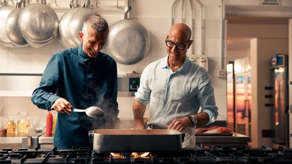 Stanley Tucci cooking with a local chef inside an authentic restaurant kitchen in Italy.