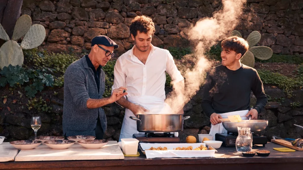 Stanley Tucci watching locals prepare traditional food over an outdoor kitchen setup.
