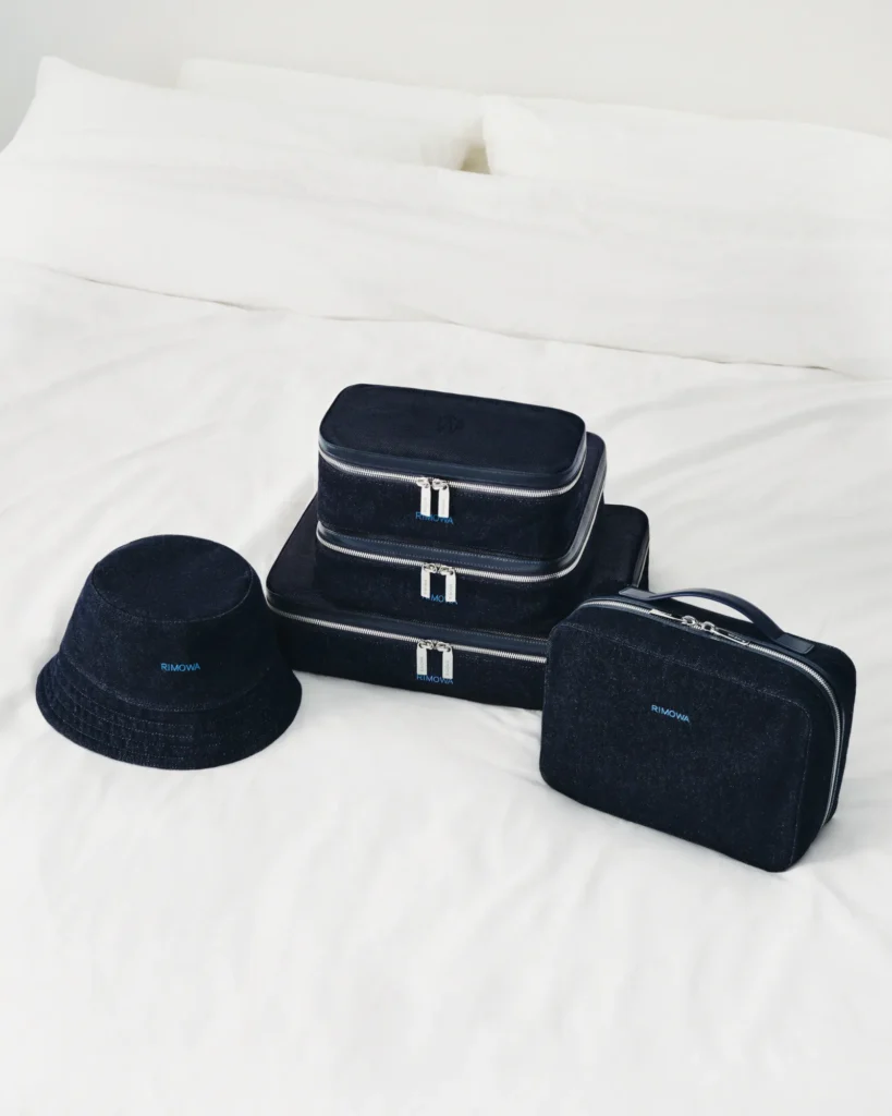 RIMOWA Ink Blue denim accessories – bucket hat, packing cubes and toiletry pouch on a bed.