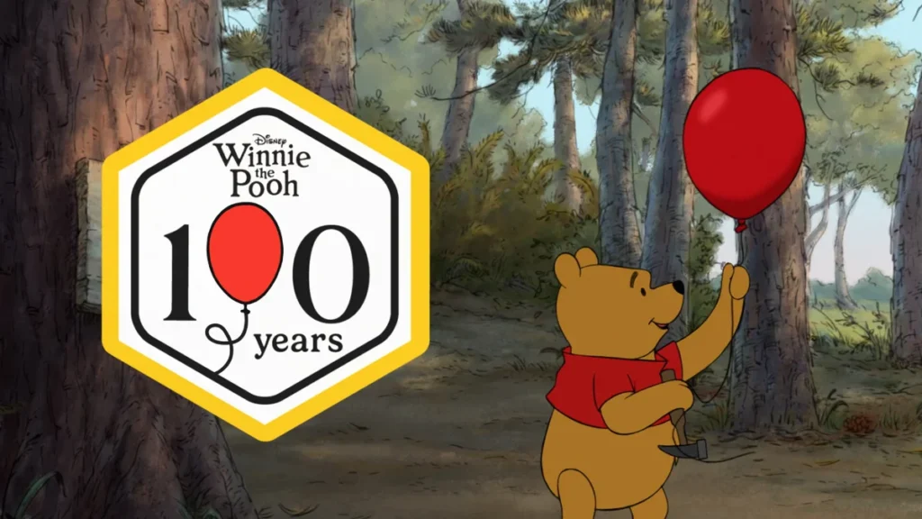 Winnie the Pooh holds a red balloon beside the Winnie the Pooh 100 Years anniversary emblem in a woodland setting.