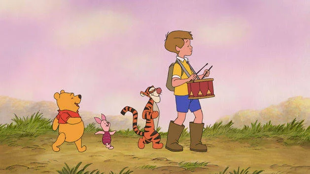 Christopher Robin carries a drum while Winnie the Pooh, Piglet, and Tigger follow him across a grassy landscape.